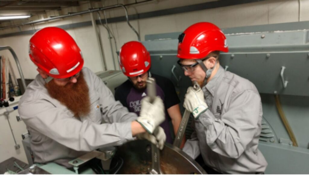 Three technicians with red helmets using wrenches on energy equipment at Laramie Community College