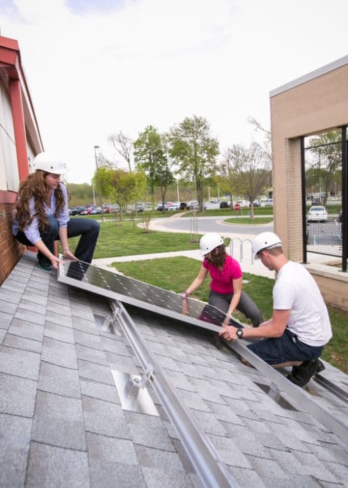 three students place a solar panel on a mock roof.